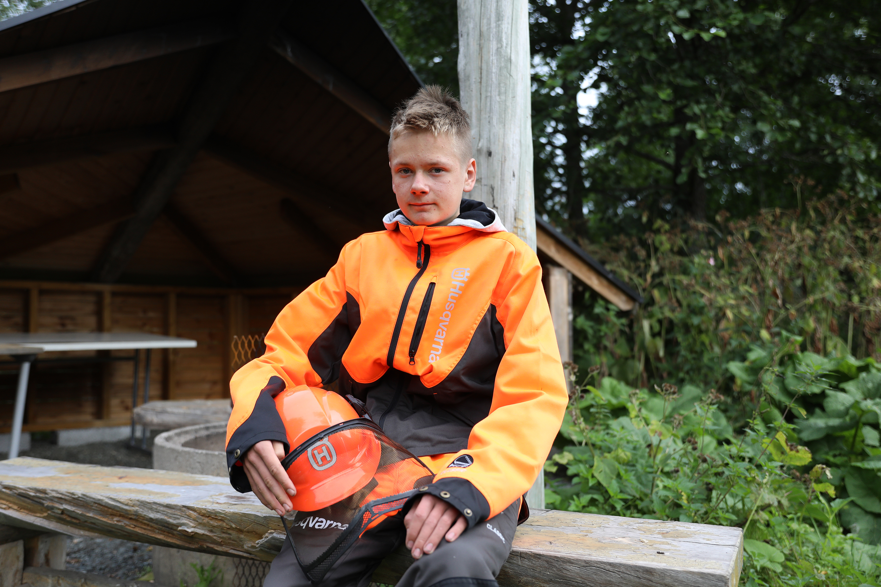 Kuvassa metsäalan opiskelija Aappo Vähäaho istuu poseeraamassa laavulla metsurinvaatteet päällään.