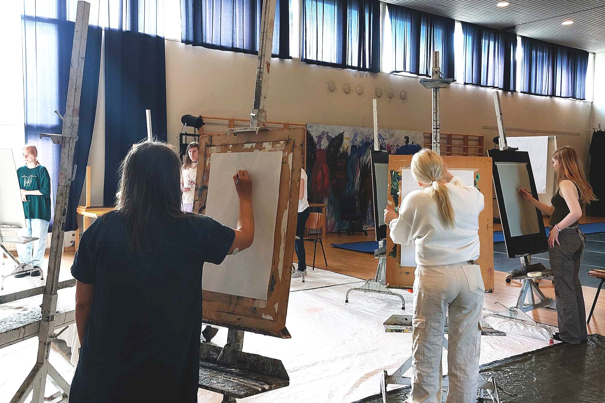 Kuvataiteen opiskelijoita aloittamassa maalaustöitään maalaustelineiden edessä liikuntasalissa.