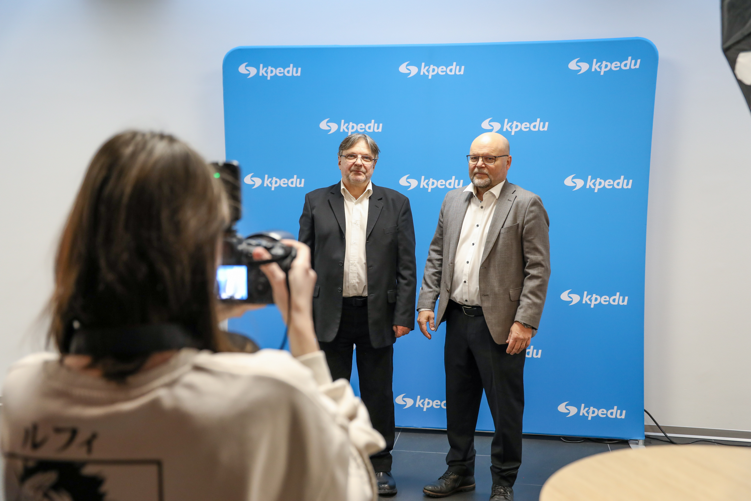 Etualall opiskelija ottaa kuvaa. Taustalla kaksi kpedulaista logoseinän edessä. 