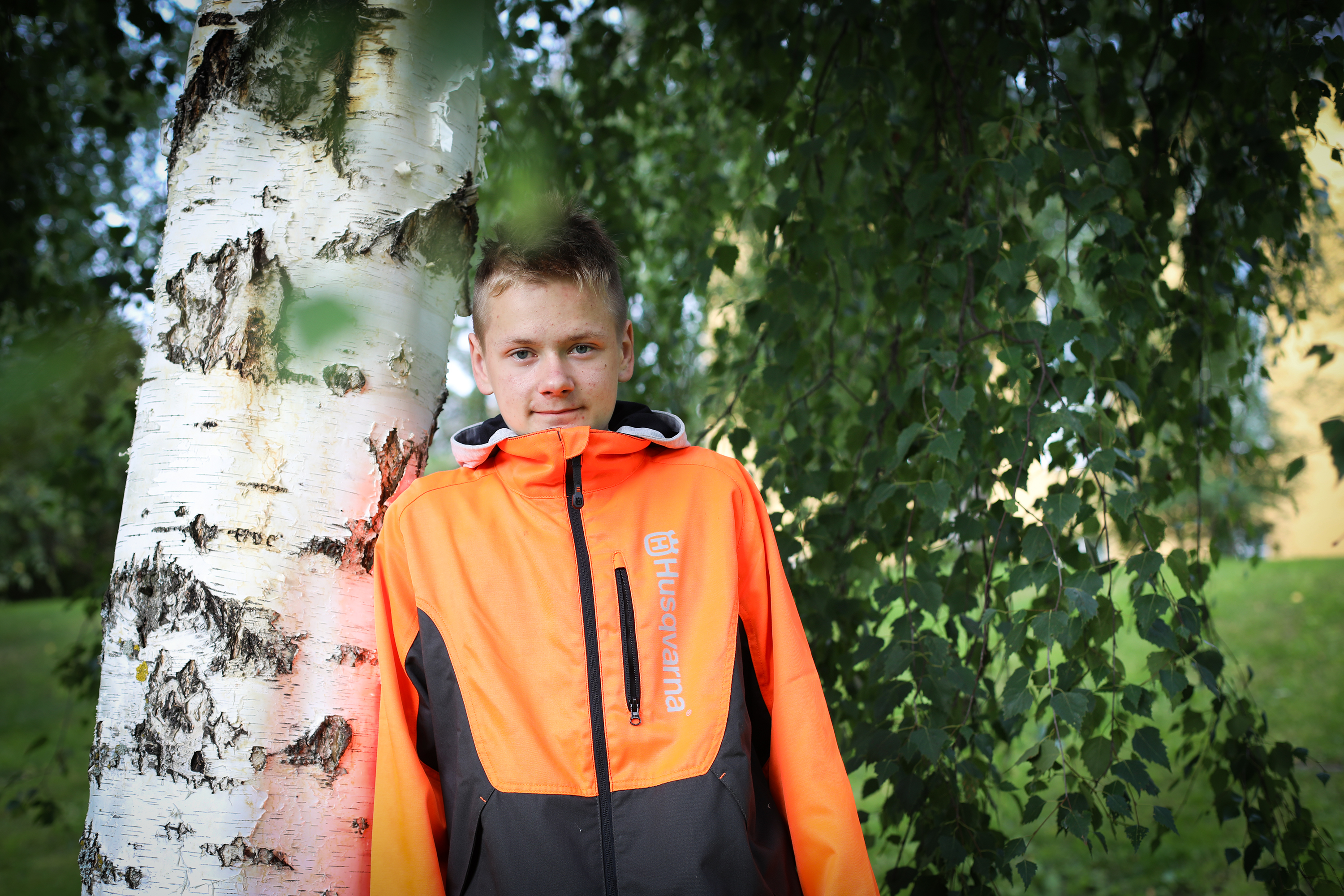 Kuvassa metsäalan opiskelija Aappo Vähäaho poseeraa nojaten koivuun metsurinkamppeet päällään.