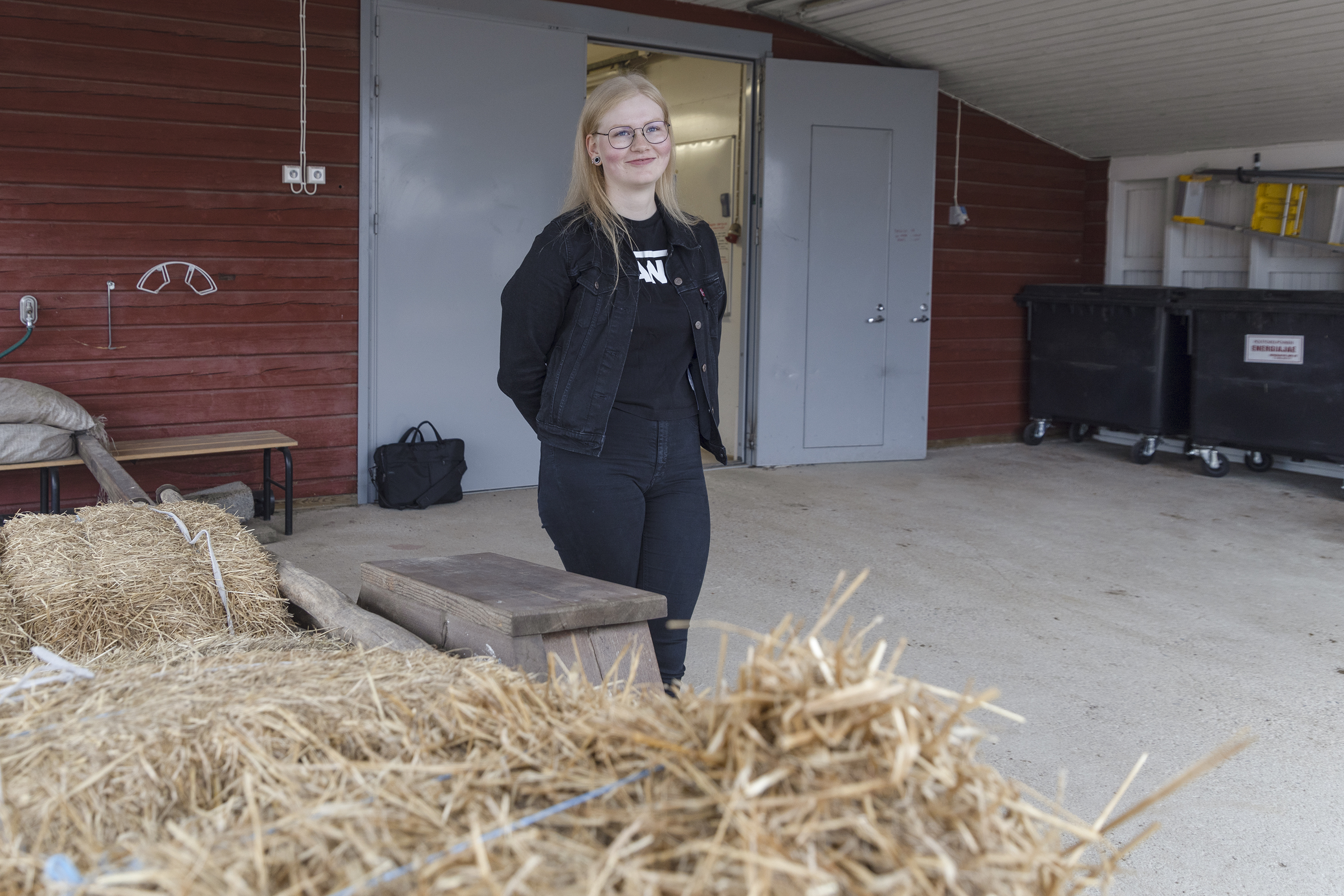 Hevosalan opiskelija Heli Haapasalo poseeraa tallin edessä seisten. Etualalla heinäpaaleja. 