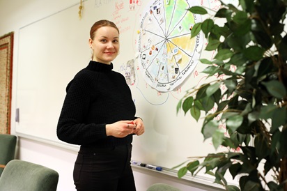 Kuvassa Lina Honkaperä poseeraa valkotaulun edessä luokassa. Etualalla on kasvi ja taululla tekstiä.