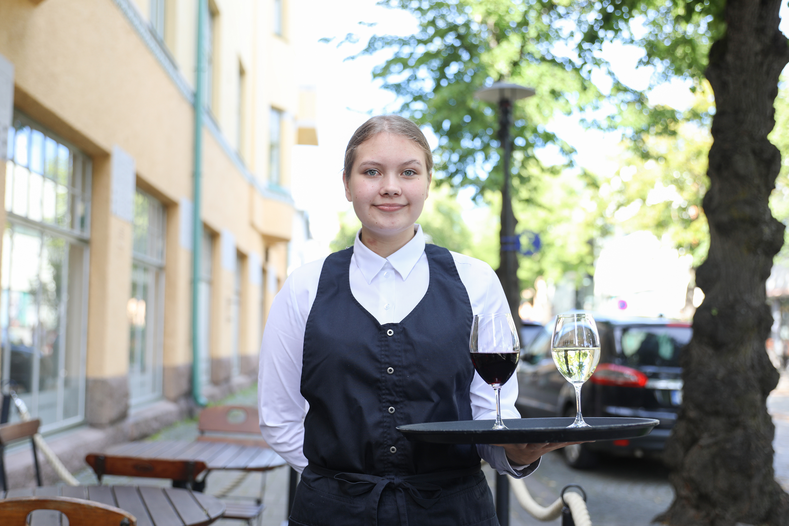 Kuvassa ravintola- cateringalan opiskelija Ronja Högland poseeraa tarjoilijan vaatteissa viinilasien kera ravintola Kokkolinnan edustalla. 