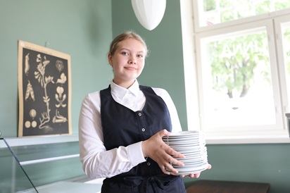 Kuvassa ravintola- cateringalan opiskelija Ronja Högland poseeraa tarjoilijan vaatteissa lautaspinon kanssa.